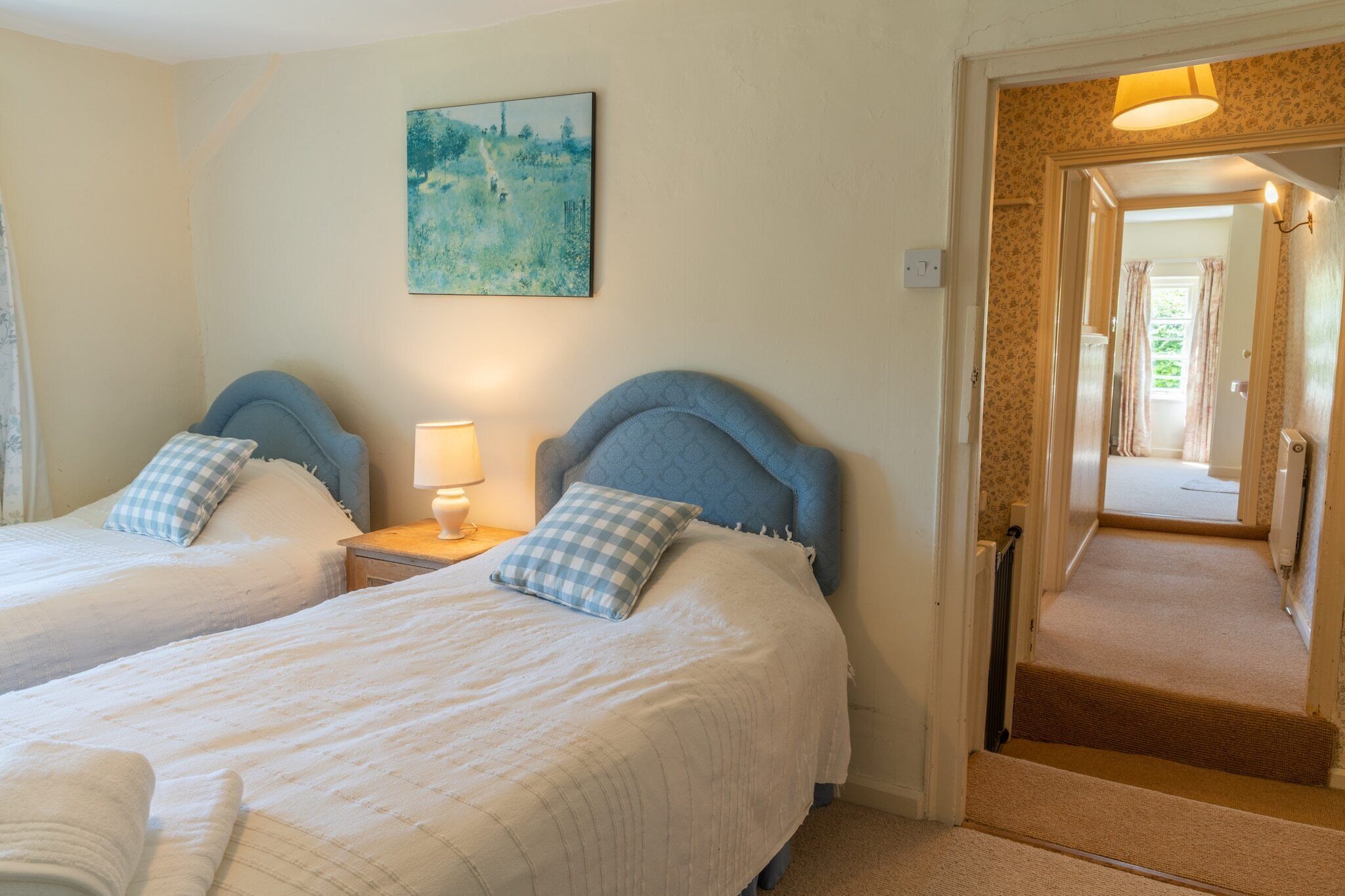 A hotel room with two beds, a painting on the wall, and a hallway leading to another room.