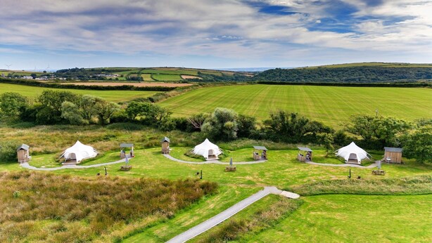 Ein Campingplatz mit mehreren weißen Zelten und Holzhütten, umgeben von grünen Feldern und Hügeln.