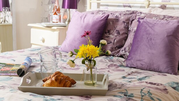 Ein ordentlich bezogenes Bett mit einer blumigen Tagesdecke, ein Tablett mit einem Croissant und einem Glas Wasser sowie eine Vase mit gelben Blumen.