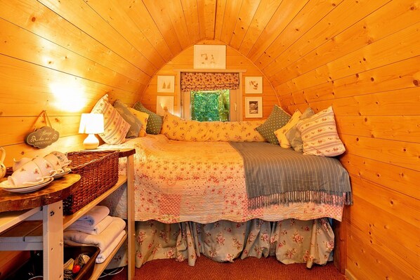 A cozy, wooden-paneled room with a bed, a bedside table with a lamp, a window with a view of greenery, and a shelf with towels and a basket.