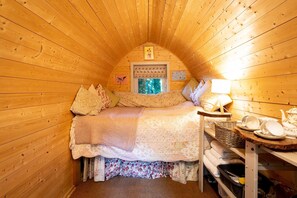 Une chambre mansardée chaleureuse avec des murs en bois, un lit recouvert d’une literie à motifs floraux, une fenêtre avec des volets et une petite table sur laquelle sont posées des assiettes.