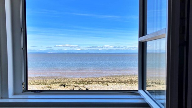 Udsigt gennem et vindue af en strand med klart himmel og roligt hav.
