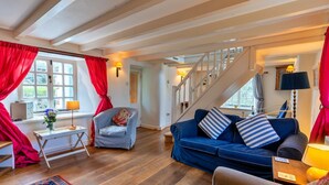 Een woonkamer met een blauwe bank, een grijze fauteuil, houten vloeren, een trap en een raam met rode gordijnen.