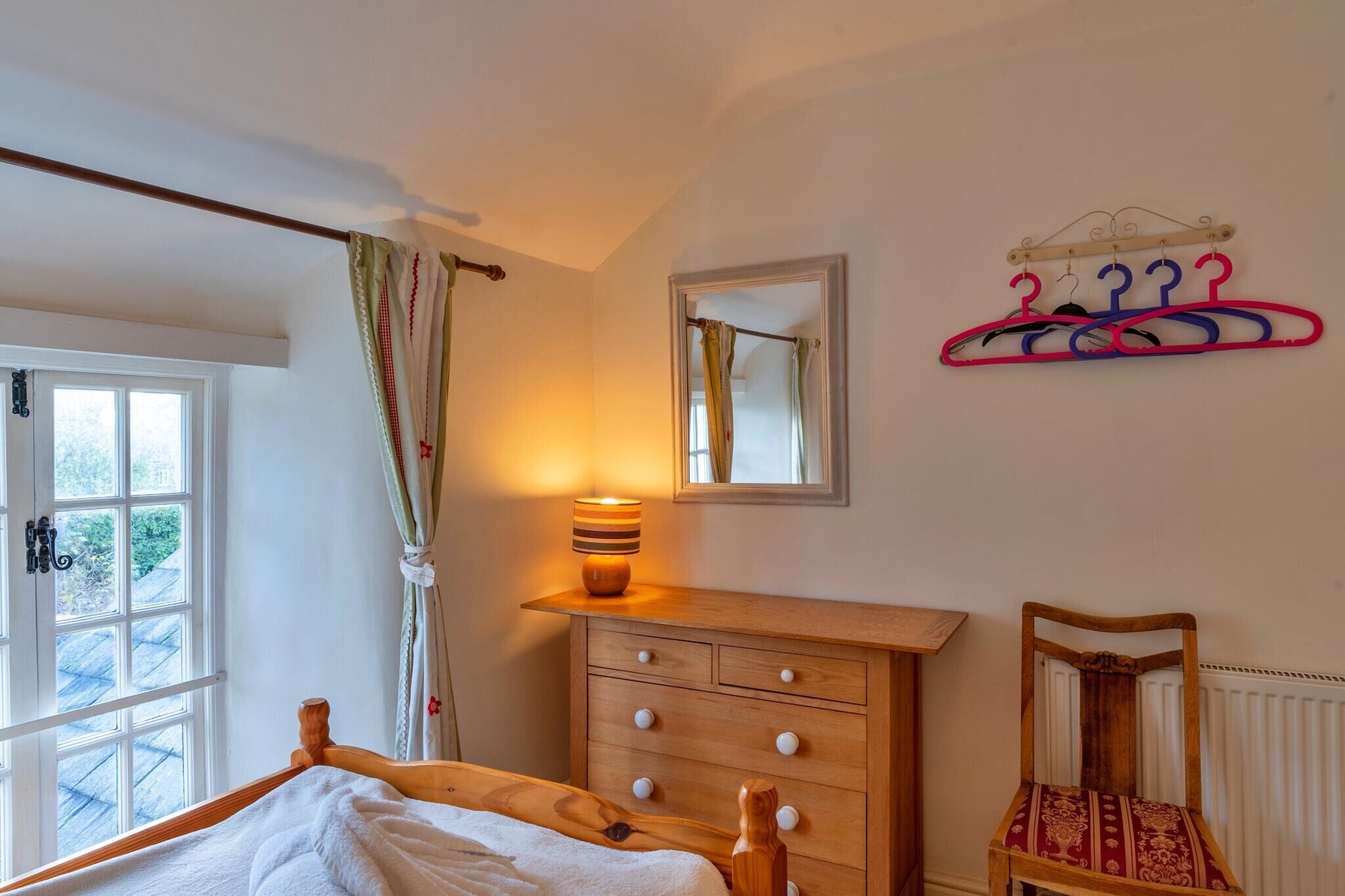A bedroom with a bed, a wooden dresser, a chair, a mirror, and a wall-mounted clothing rack.