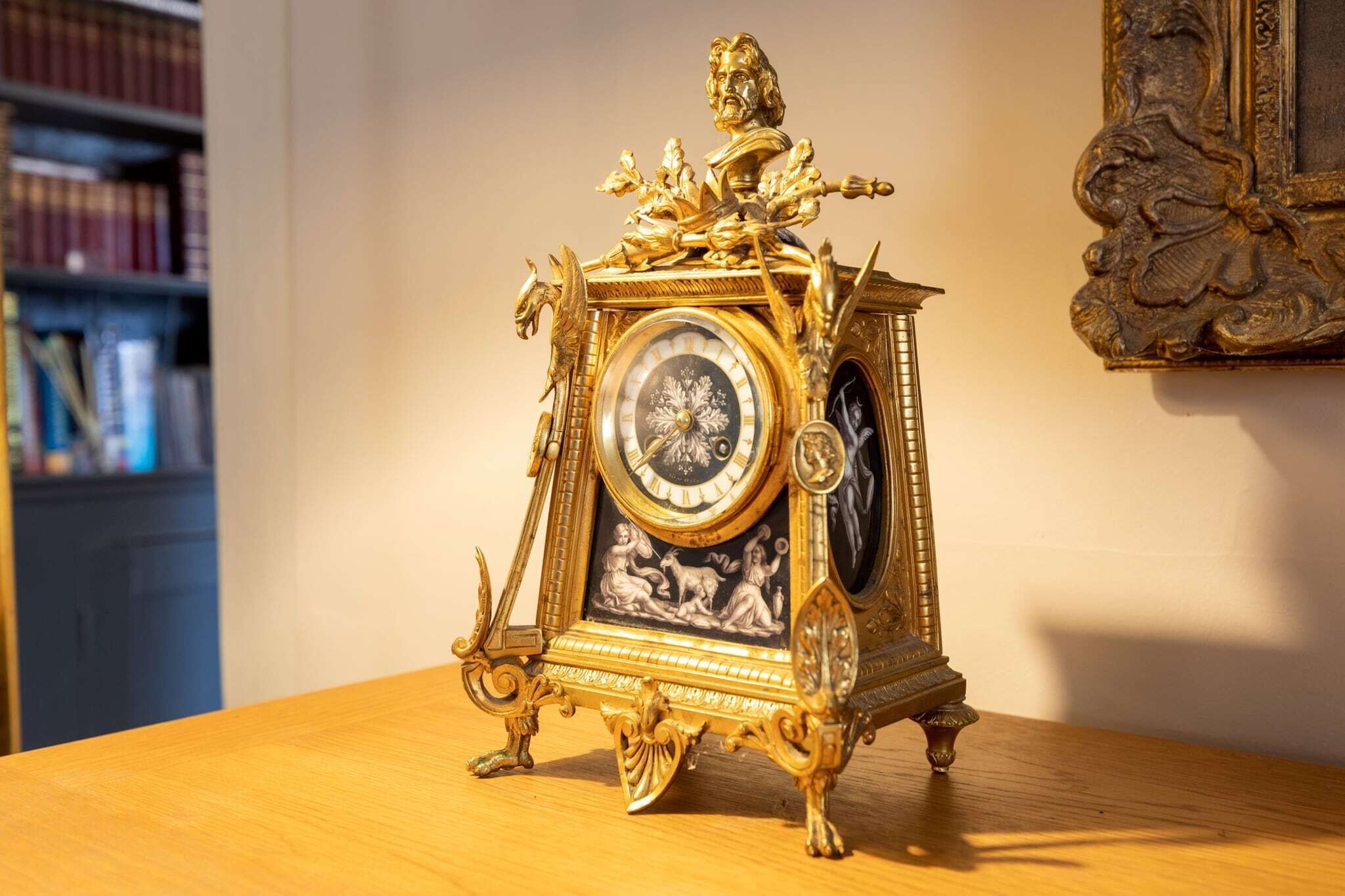 Een gouden, sierlijk uurwerk met een gedetailleerde wijzerplaat en een beeldje bovenop, staand op een houten tafel.