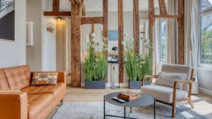 Ein Wohnzimmer mit einer Couch, einem Sessel und einem Couchtisch, Holzbalken, Topfpflanzen und Blick ins Freie.