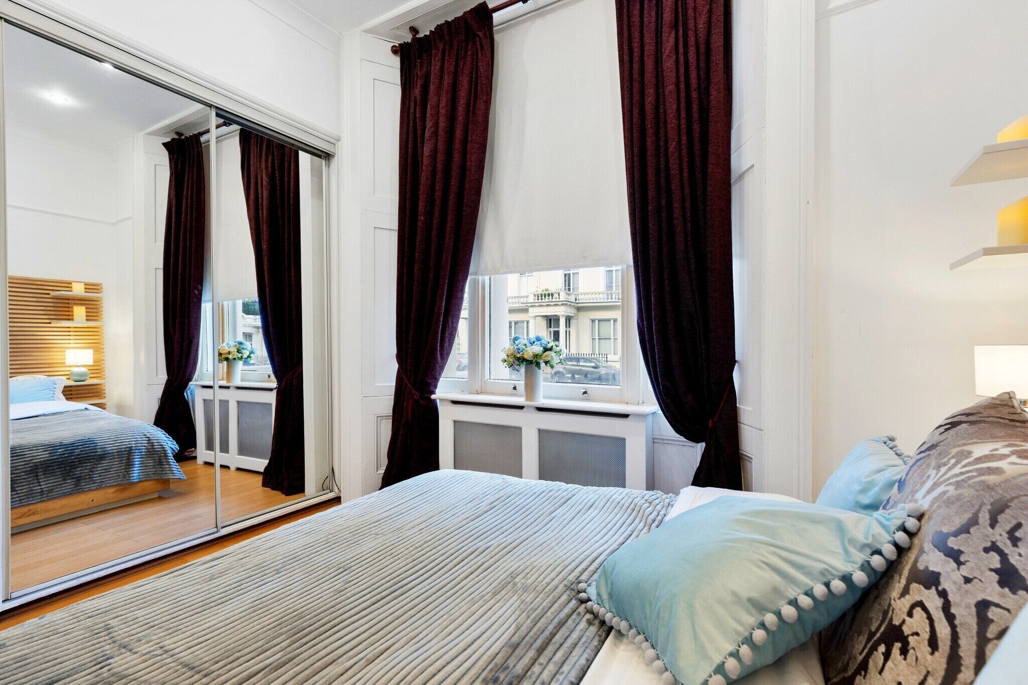 Un dormitorio con cama, ventana con cortinas, mesita de noche con un florero y vista a la calle.