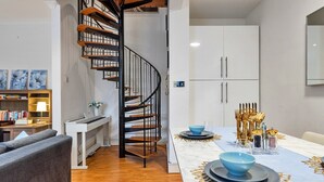 Un salon moderne doté d’un escalier en colimaçon, d’un coin repas avec une table blanche dressée pour deux personnes et d’une bibliothèque remplie de livres et d’objets décoratifs.