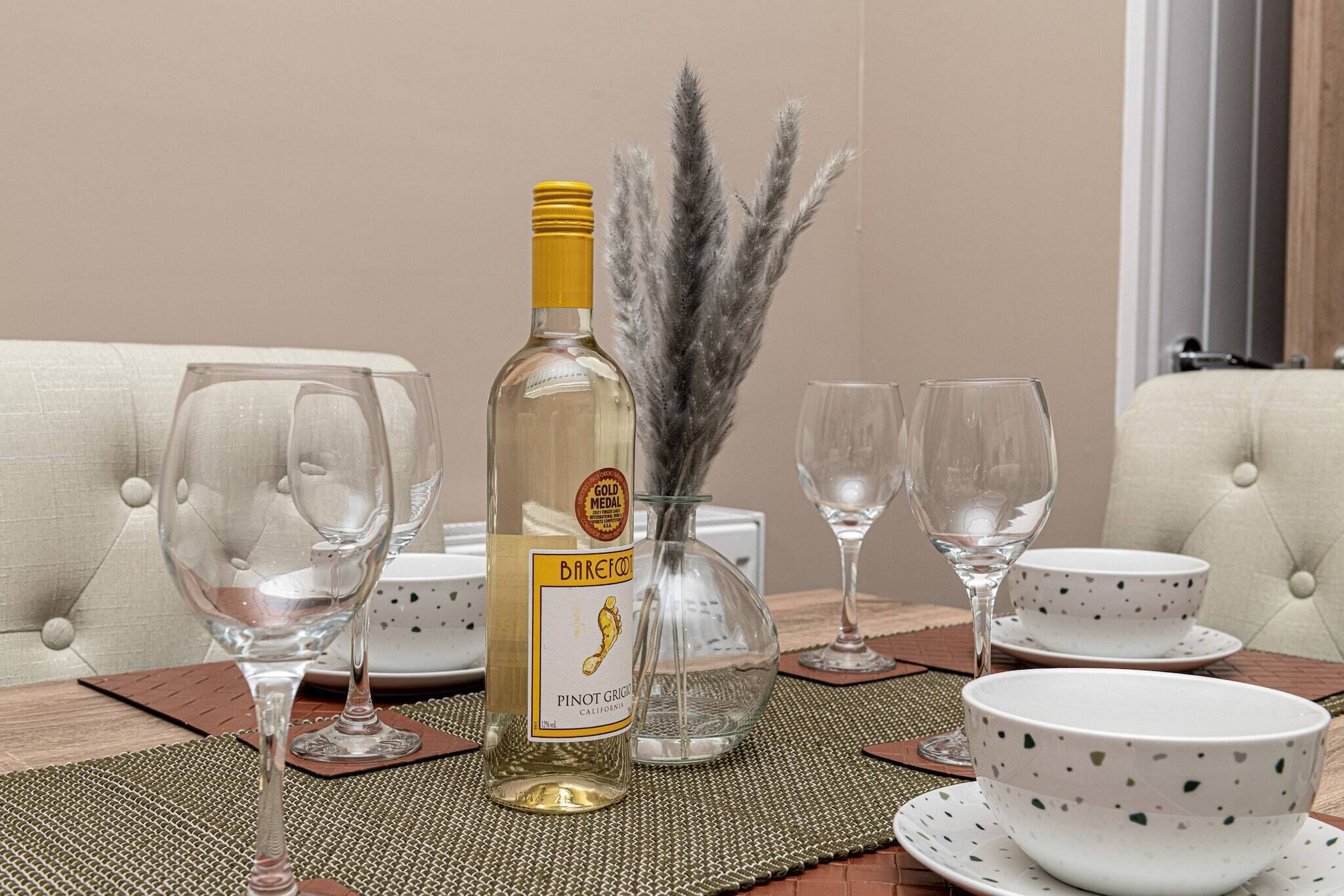 A dining table with a bottle of Barefoot Pinot Grigio, two wine glasses, a vase with dried pampas grass, and a set of white plates and bowls.