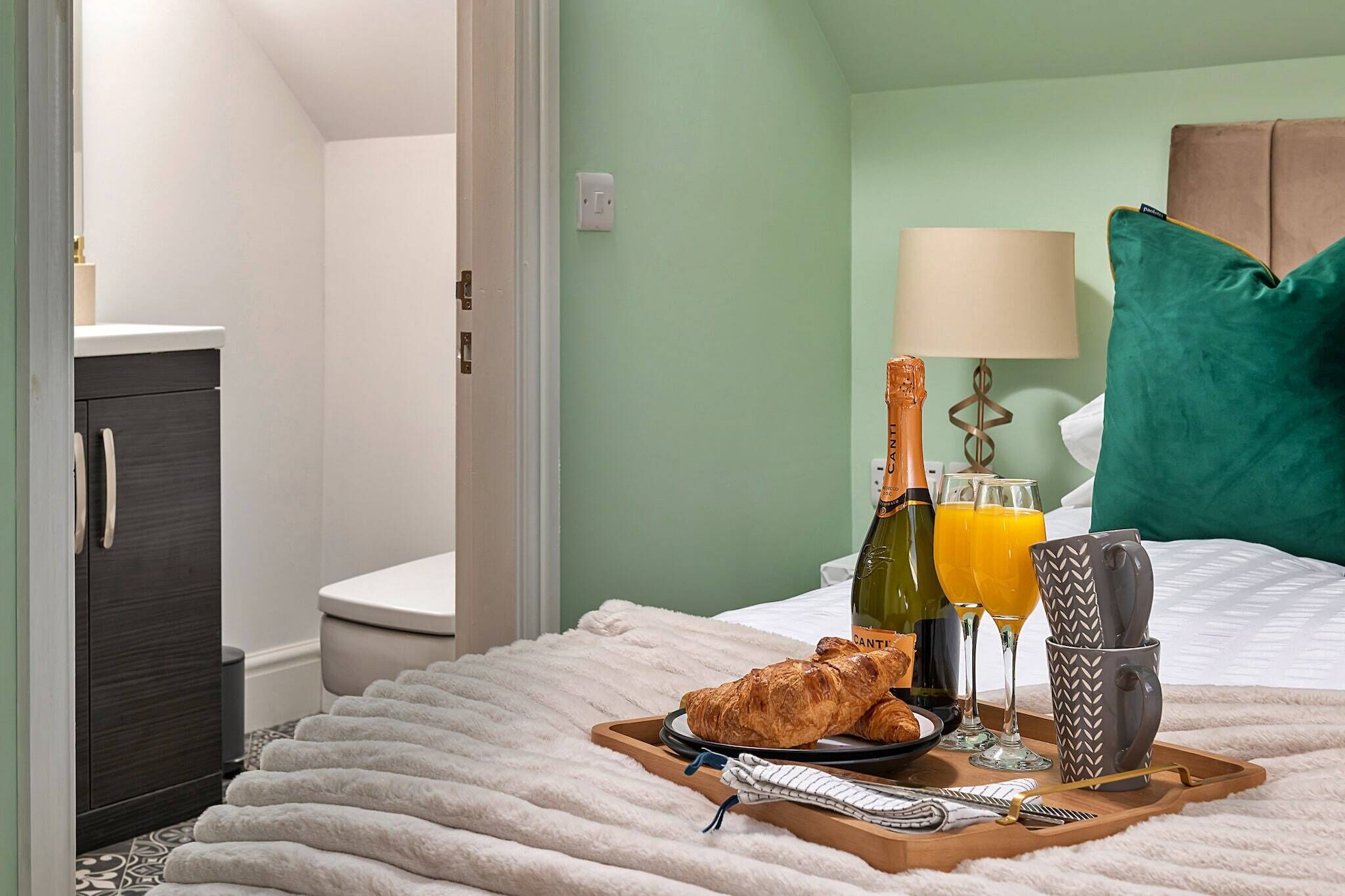 A hotel room with a bed, a bedside table with a lamp, a green pillow, a tray with croissants, a bottle of champagne, and glasses of orange juice.