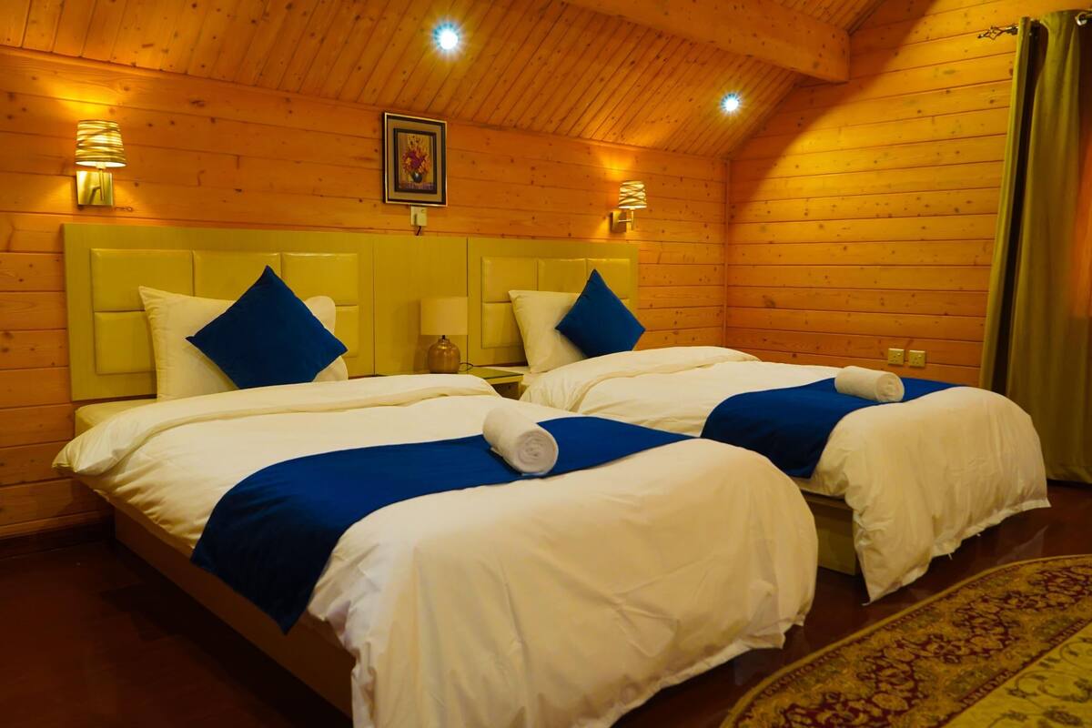 Dormitorio de cabaña de madera con dos camas, cada una con ropa de cama blanca y detalles azules, y techo de madera.