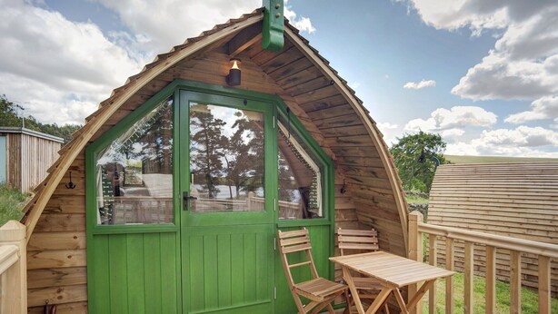 Una cabaña de madera con puerta verde, una mesita y una silla en una terraza de madera.