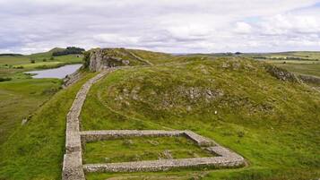 A stone structure with a rectangular courtyard, surrounded by grassy hills and a body of water in the background.