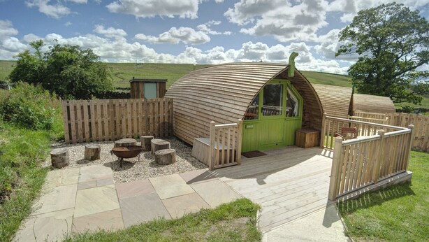 Una cabaña de madera con puerta verde, una terraza y una zona con fogata.