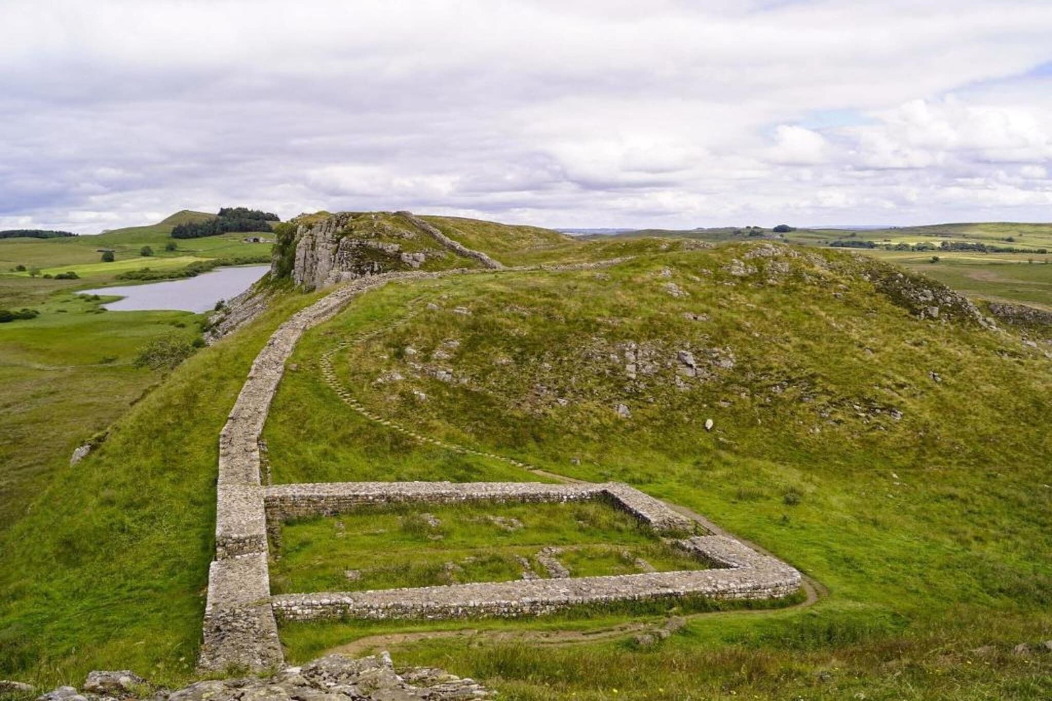 A stone structure with a rectangular layout, surrounded by grassy hills and a body of water in the background.