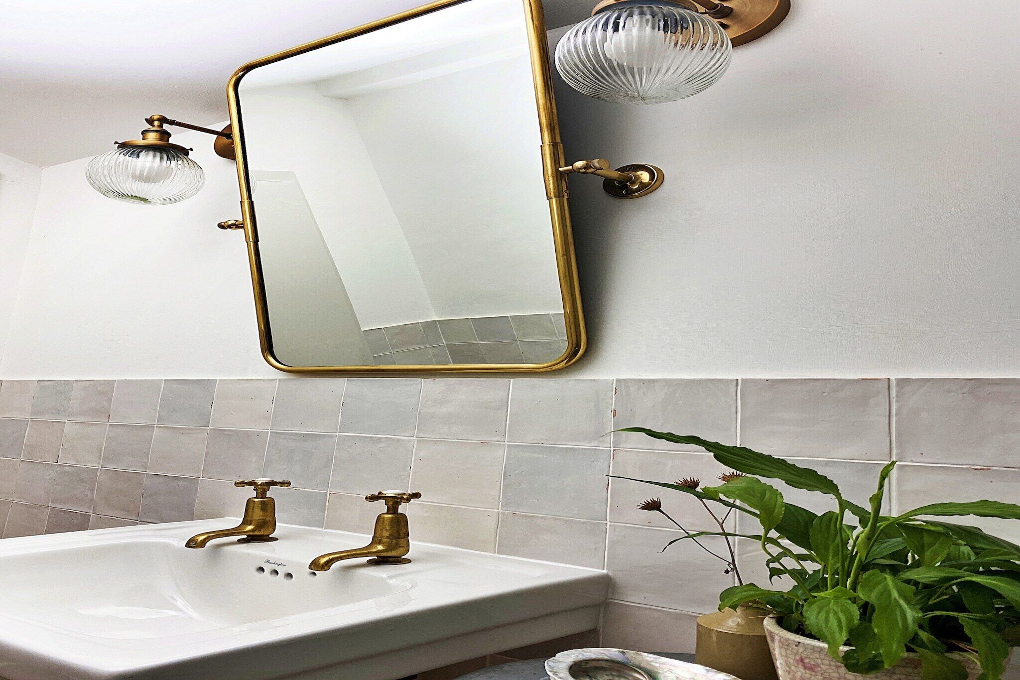 A bathroom with a white sink, brass faucets, and a large mirror with a gold frame. There are two wall-mounted lights and a potted plant.