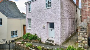Un edificio de piedra rosa con puerta y ventanas blancas, un pequeño jardín y un muro de piedra.