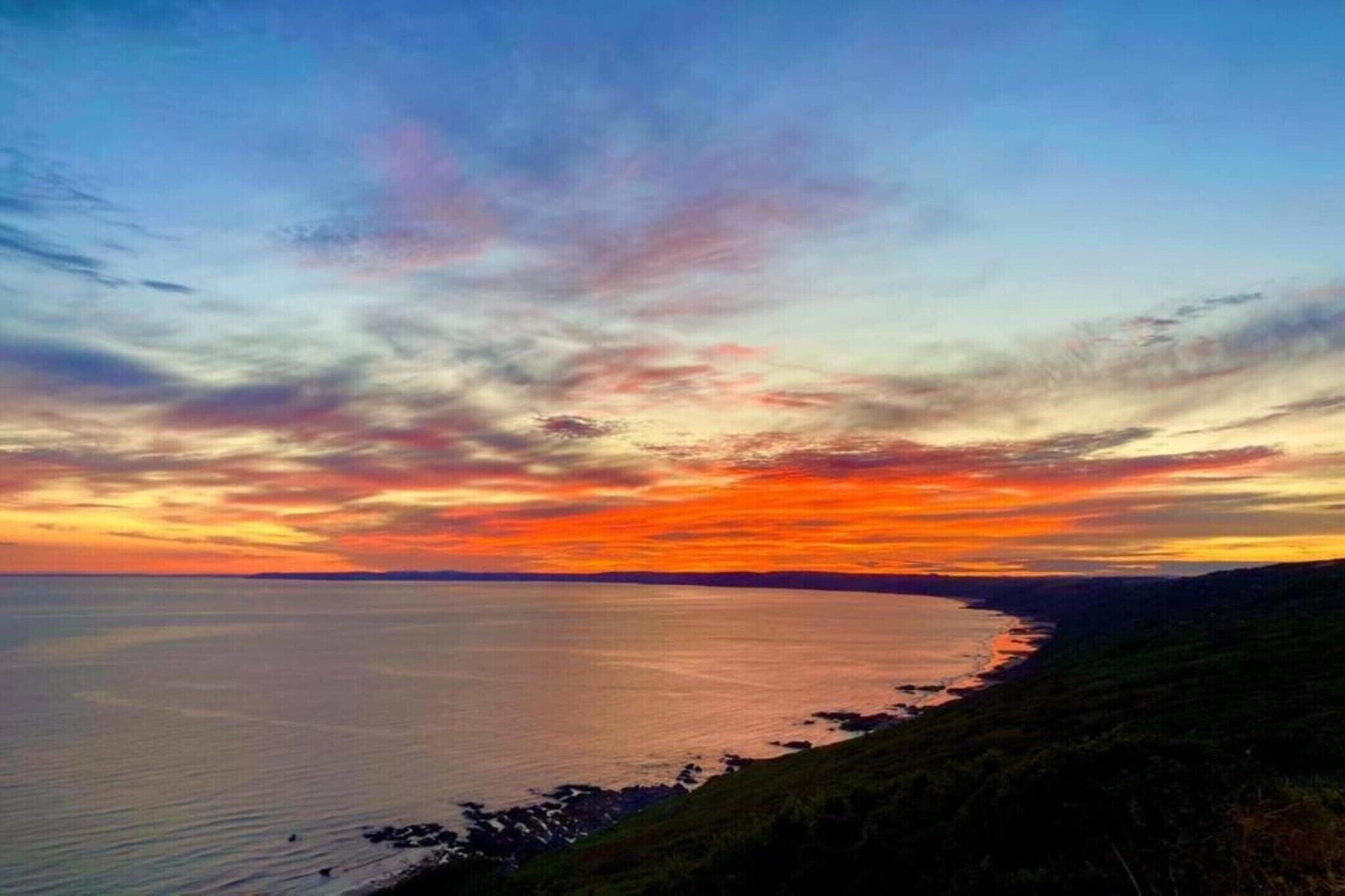A coastal landscape during sunset with a clear view of the sea and distant shoreline.