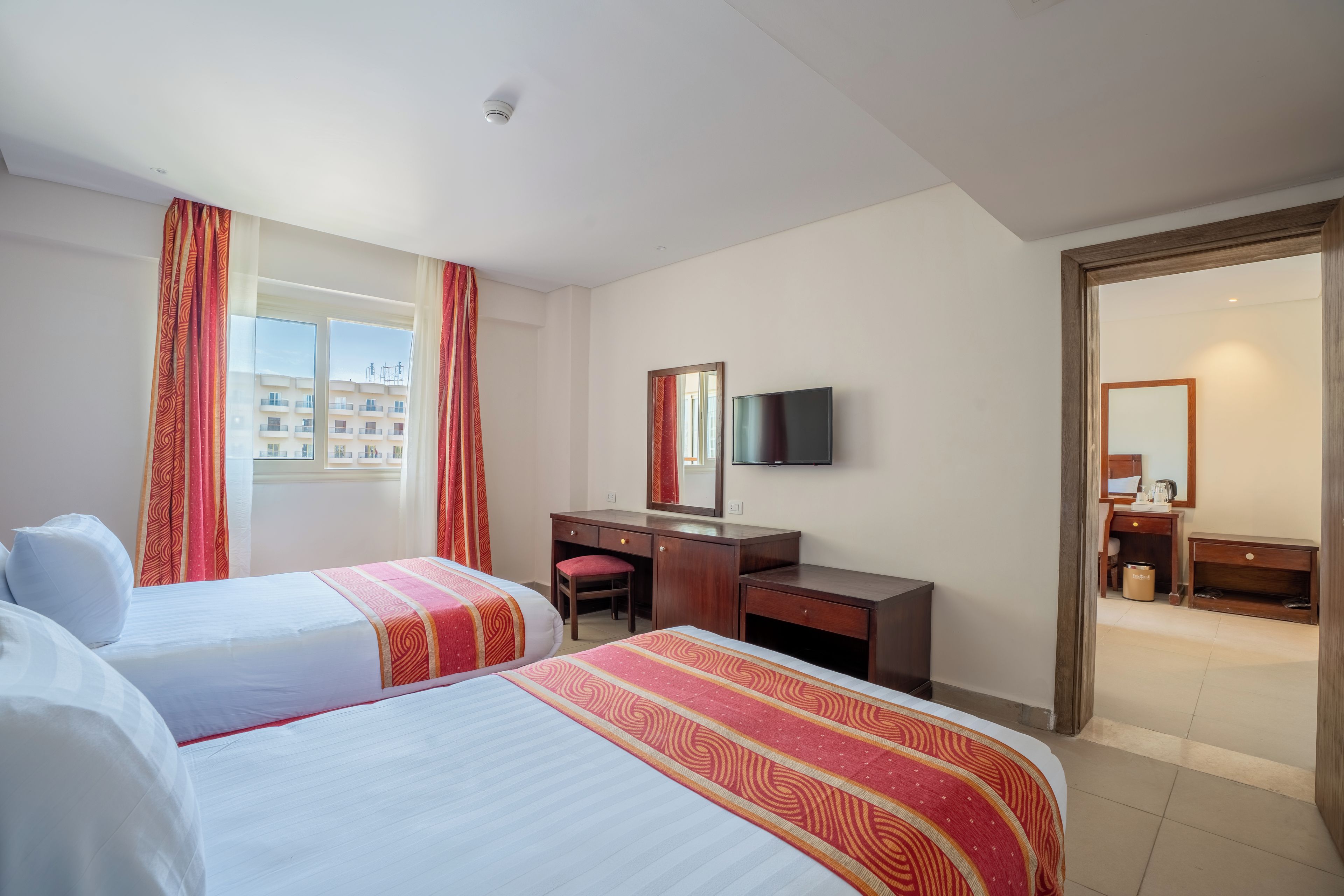 Habitación de hotel con dos camas, un escritorio, una silla, un televisor y un baño visible a través de una puerta abierta.