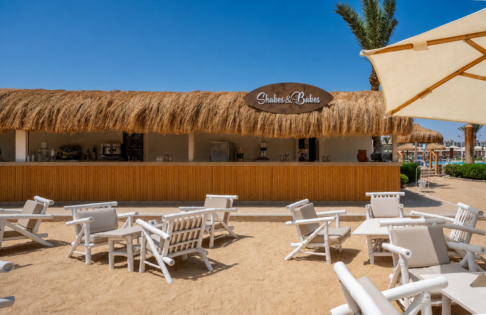 Un bar frente al mar llamado 'Shaker & Bakes' con techo de paja, sillones blancos y una zona de playa arenosa.