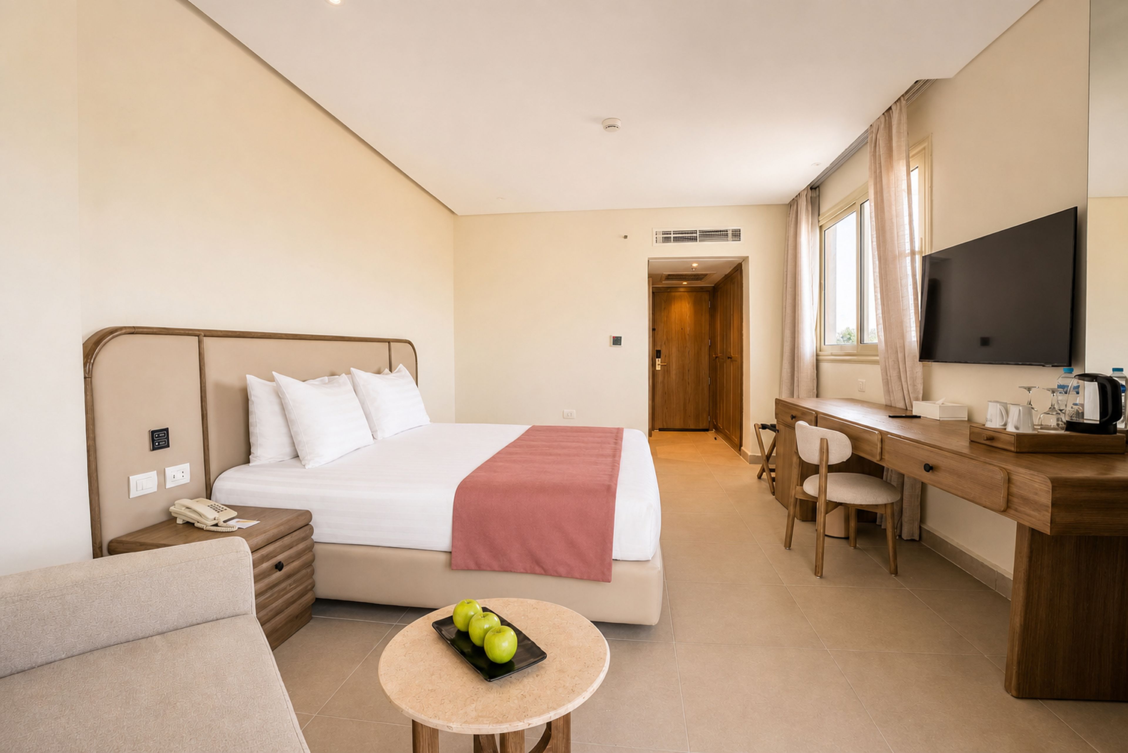 A hotel room with a bed, a desk, a chair, a small table with apples, and a television.