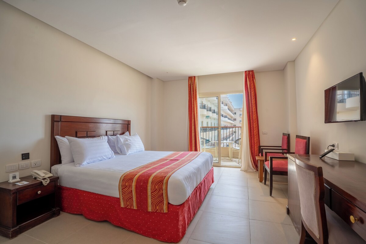 Habitación de hotel con cama, mesita de noche, escritorio, silla y vista a un edificio a través de una ventana.