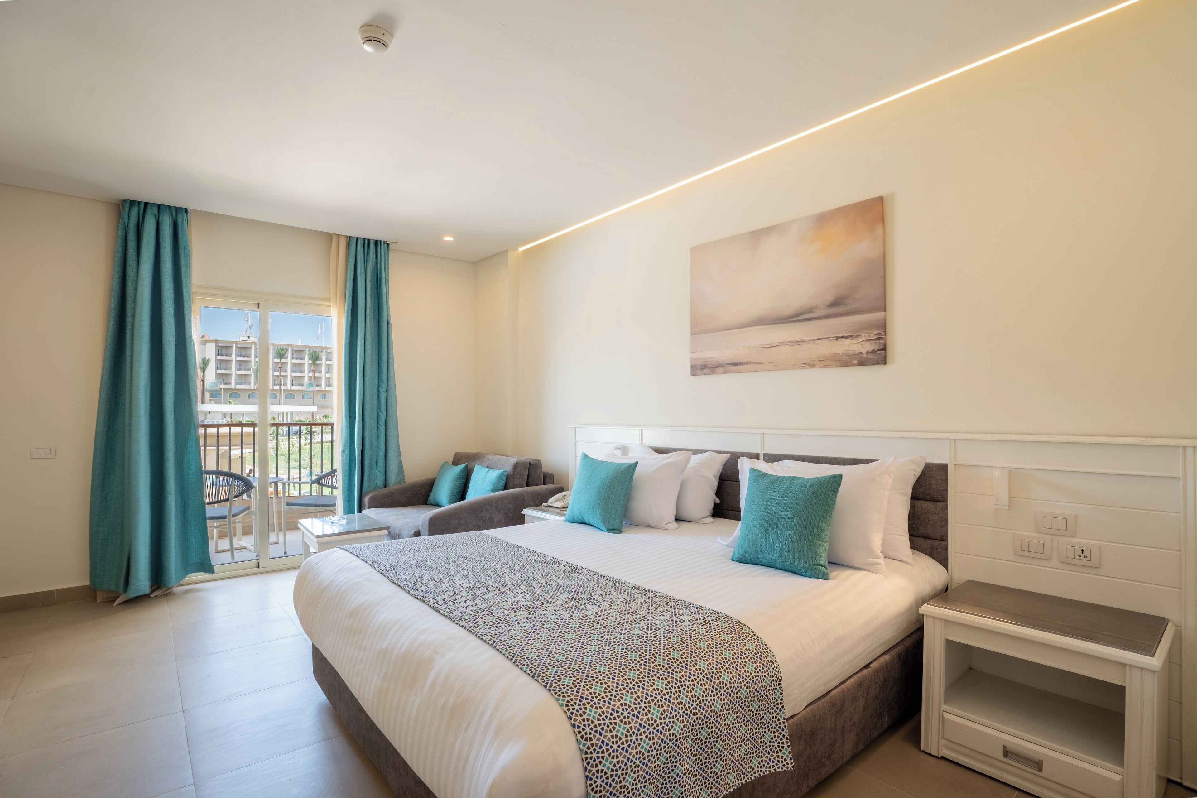 Quarto de hotel com cama, mesa de cabeceira, pintura, varanda com cadeiras e cortinas.