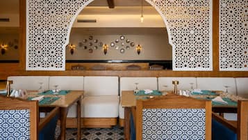 A restaurant interior with wooden tables, cushioned seating, decorative wall panels, and wall-mounted light fixtures.