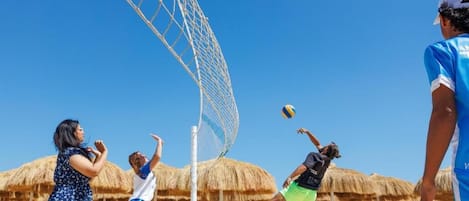A beach volleyball game with players in casual beach attire, a volleyball net, and thatched umbrellas in the background.