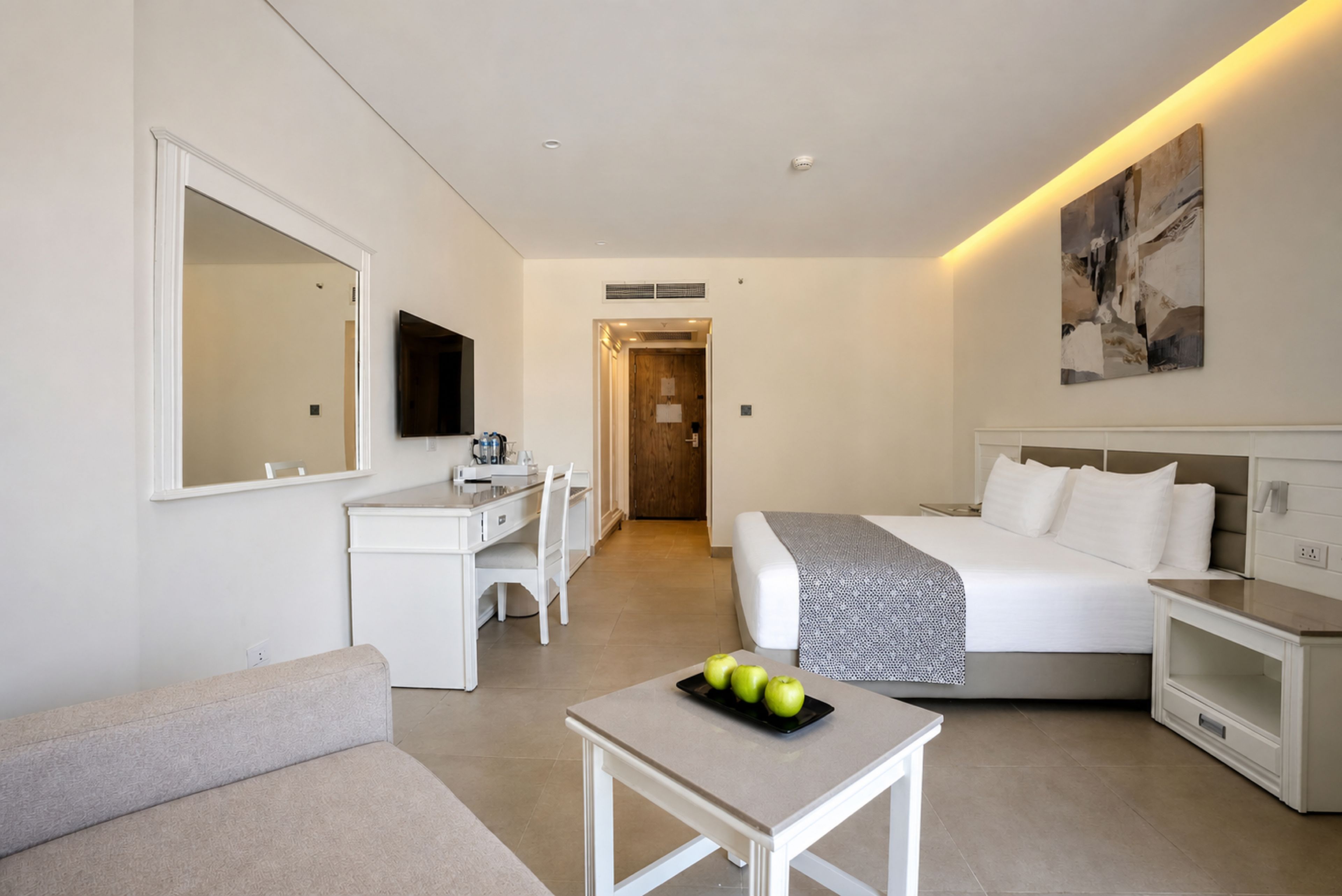 Una camera d'albergo con un letto, una scrivania, una sedia, un tavolino con delle mele, una televisione e opere d'arte alle pareti.