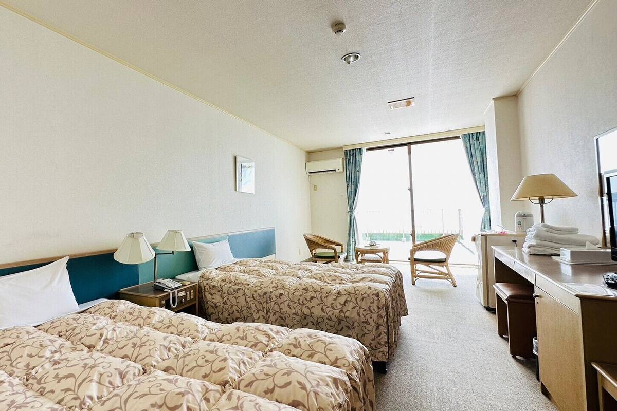 A hotel room with two beds, a desk, a chair, a small table, and a balcony with a view.