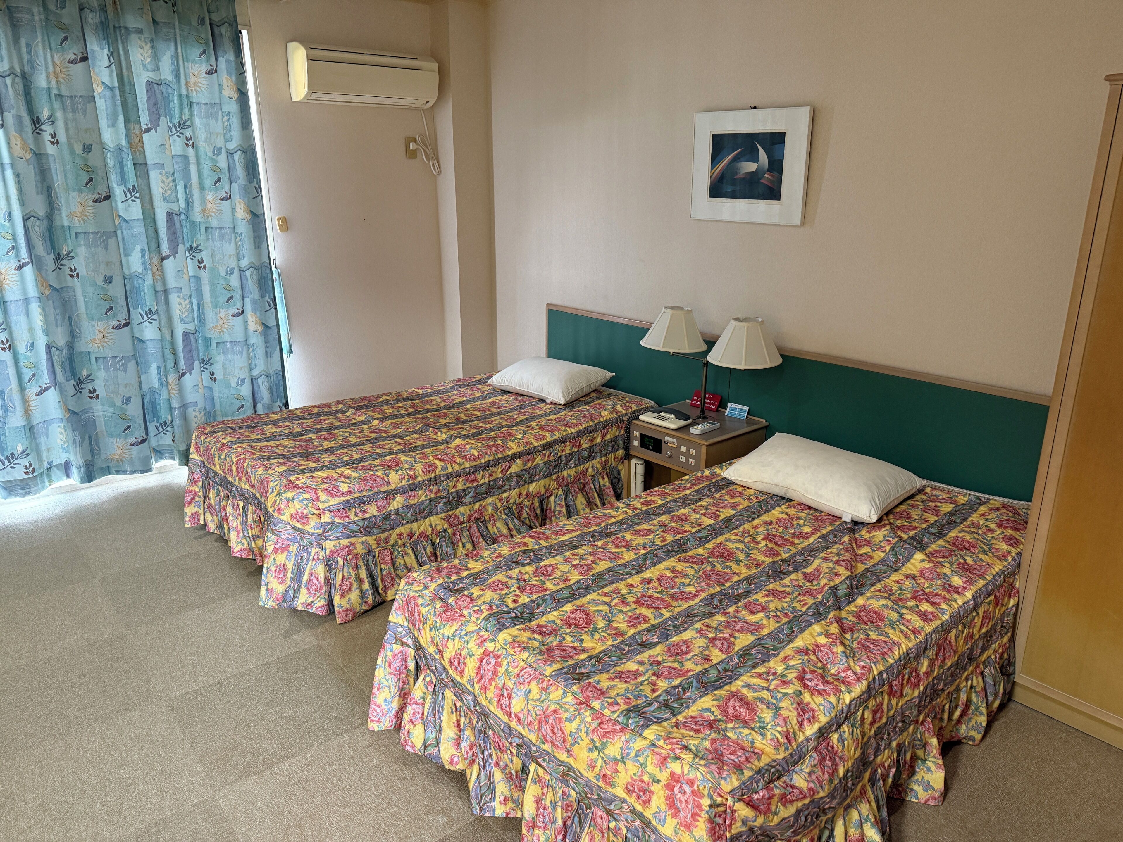 Een hotelkamer met twee bedden, elk voorzien van bloemenbedrukking, een nachtkastje met lampen, een wandairconditioner en een ingelijst schilderij aan de muur.
