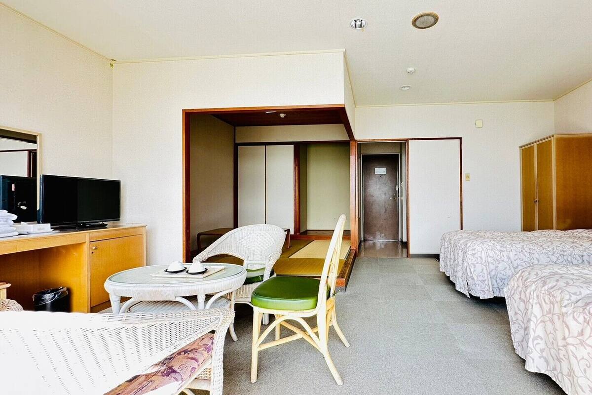 A hotel room with a bed, a desk with a TV, a dining table with chairs, and a wardrobe.