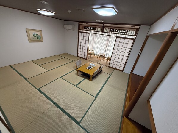Ein Raum mit Tatami-Matten, einem niedrigen Tisch, Stühlen, einer Schiebetür mit Gittermuster und einem gerahmten Bild an der Wand.