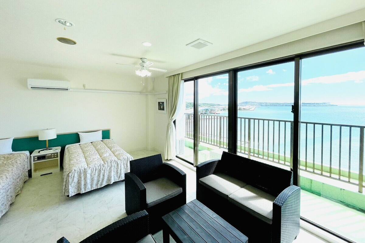 A hotel room with a bed, two bedside tables, a desk, a chair, a sofa, and a balcony with a view of the sea.