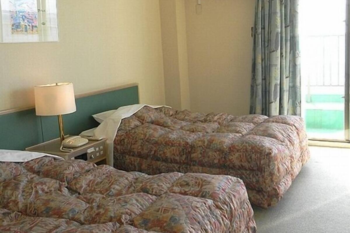 A hotel room with two beds, a nightstand, a lamp, an air conditioning unit, and a window with curtains.