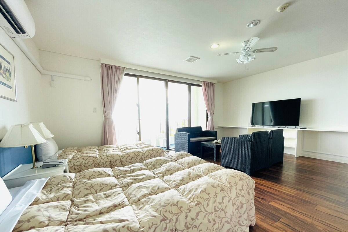 A hotel room with a bed, a desk with a lamp, a TV, a ceiling fan, and a window with curtains.