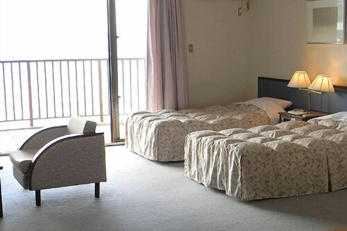 A hotel room with two beds, a chair, an air conditioning unit, a lamp, and a view of the outside.