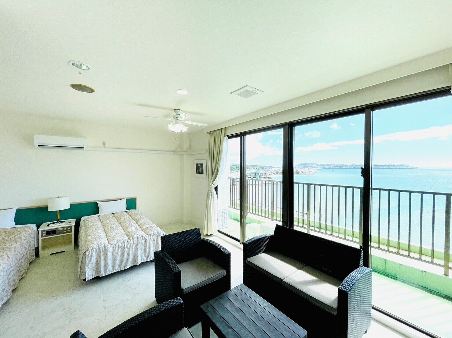 Habitación de hotel con cama, dos mesitas de noche, ventilador de techo, ventana grande con vista al mar y balcón con mobiliario exterior.