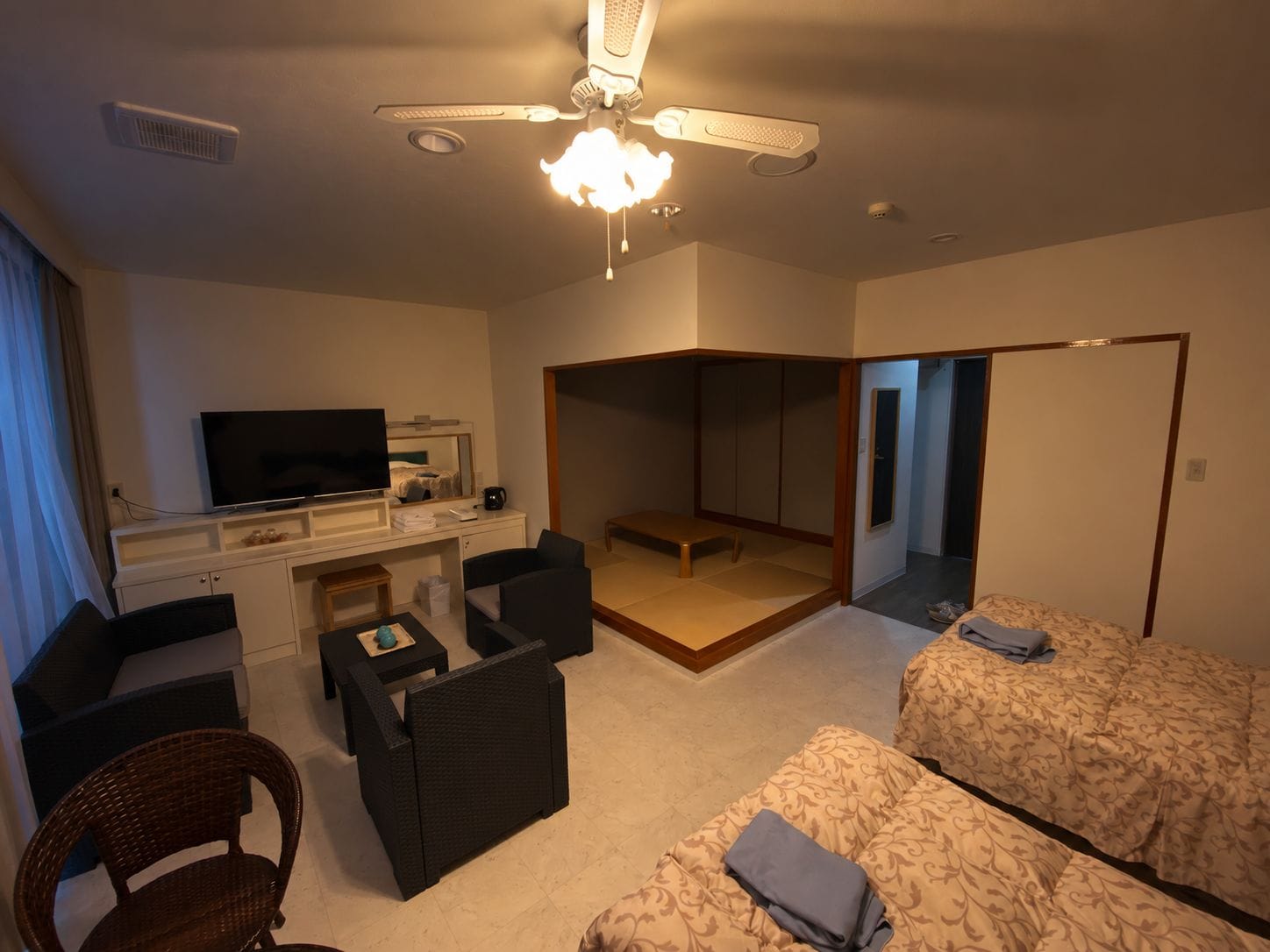 A hotel room with a bed, a desk with a TV, a chair, a sofa, and a ceiling fan.