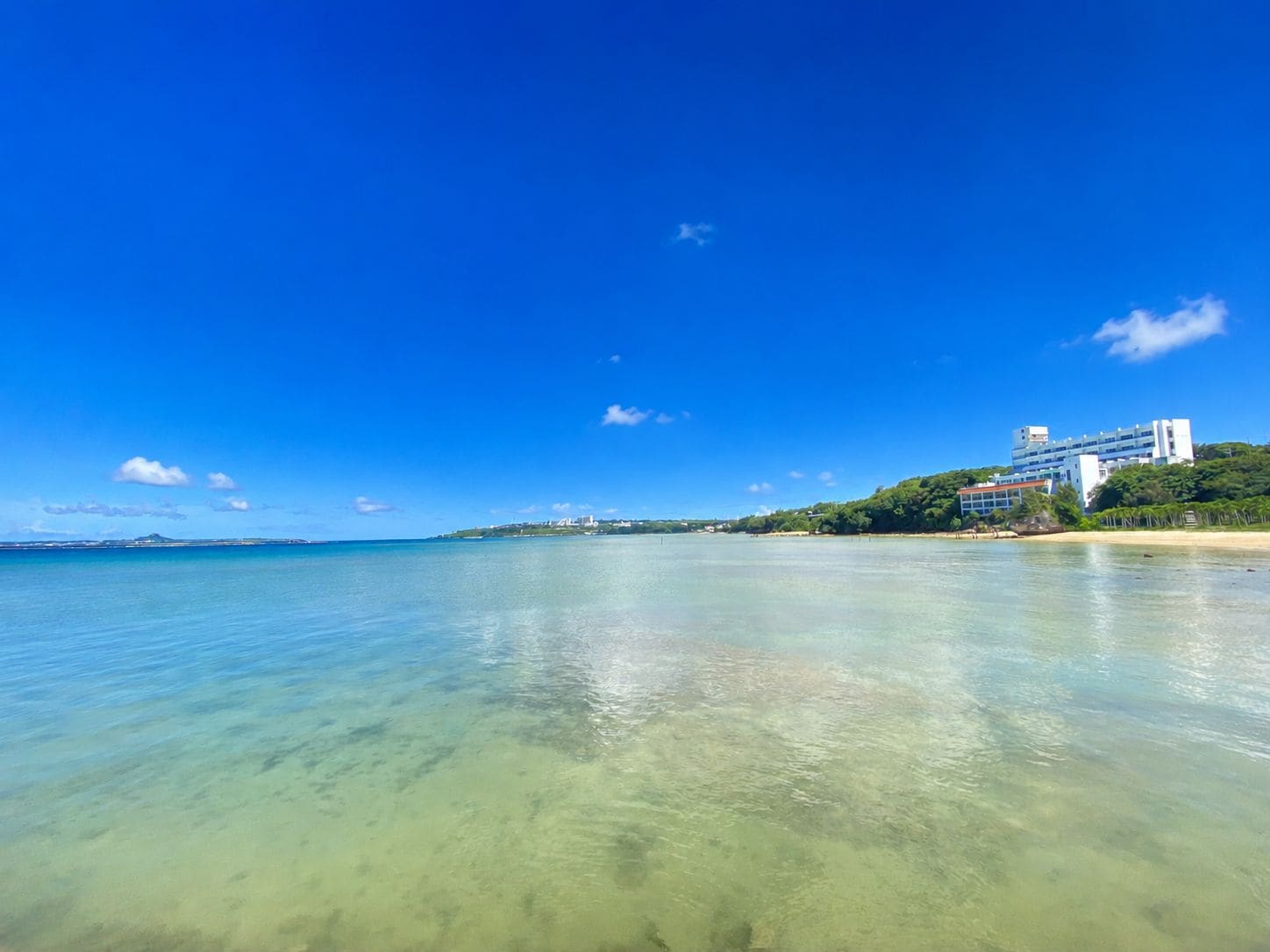 A coastal view with clear water, a beach, and a building with multiple floors.