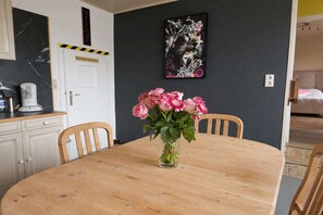 Un comedor con una mesa de madera, sillas, un jarrón de flores rosadas, una cafetera y un cuadro en la pared.
