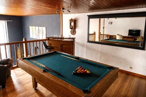 Une table de billard avec des billes, une horloge et un téléviseur, dans une pièce au plancher en bois et dotée d’une rambarde.