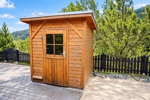 A wooden cabin with a single door and window, surrounded by a black fence and trees.