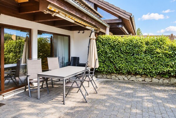 An outdoor patio with a table and chairs, a covered seating area, and a stone wall with a hedge.