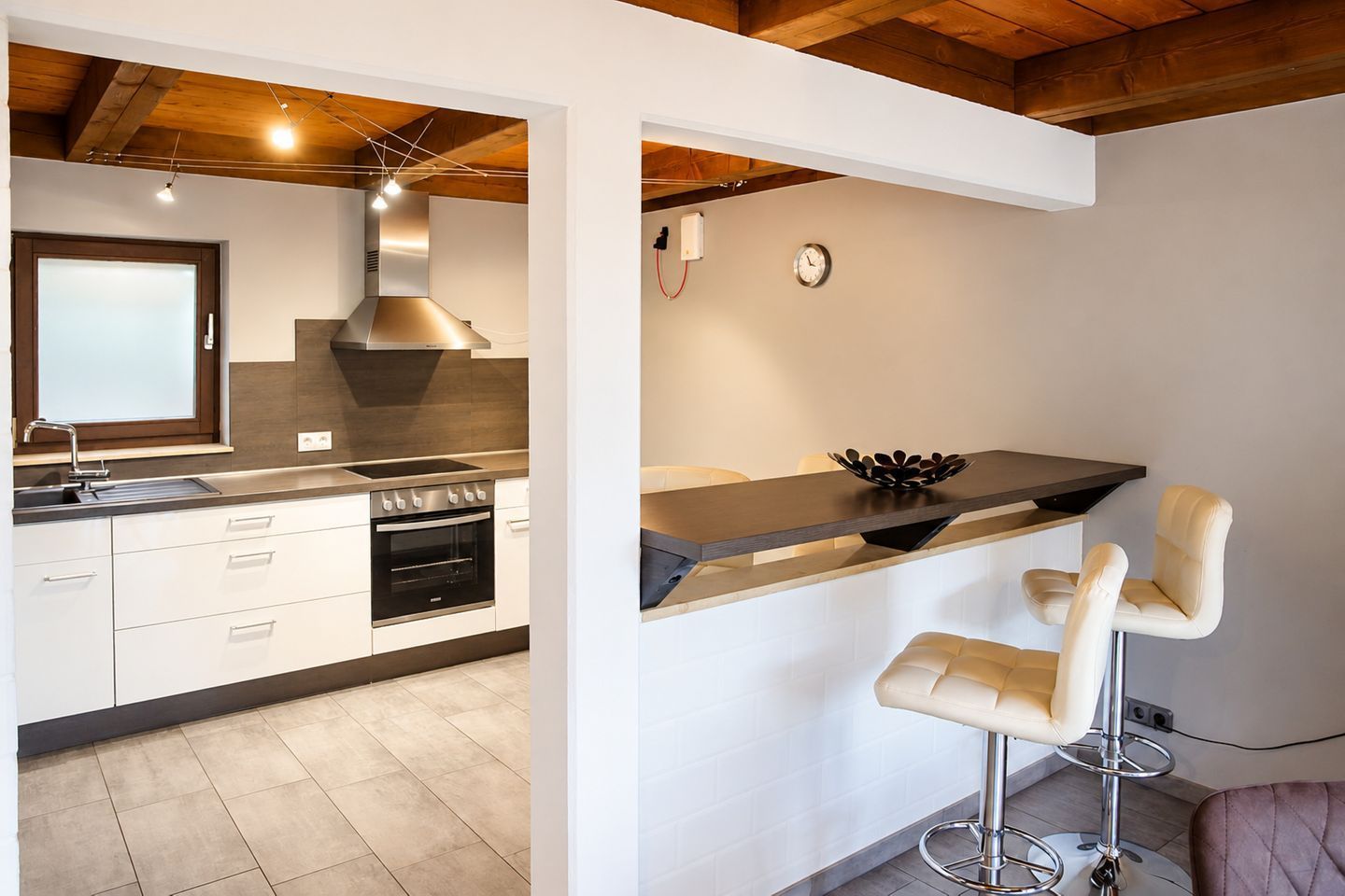 Una cocina moderna con horno empotrado, un lavamanos y una estufa. Hay una zona de bar con taburetes y un reloj en la pared.
