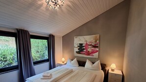A neatly made bed with towels, a bedside table with a lamp, a painting on the wall, and a window with curtains.