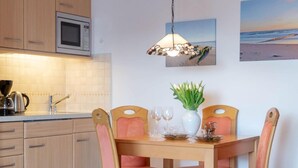A modern kitchen with wooden cabinets, a microwave, and a dining area with a glass table and pink chairs. - Coffee, cake and the smell of the coast (Sylt)