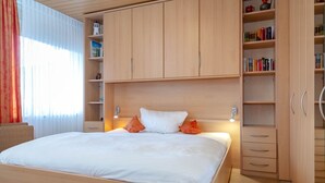 A neatly arranged bedroom with a large bed, wooden wardrobe, and built-in shelving with books and decorative items. - Coffee, cake and the smell of the coast (Sylt)