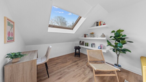 Una habitación con techo inclinado, tragaluz, suelo de madera, una silla blanca, una silla de mimbre, una pequeña mesa, una planta en maceta y una estantería con libros y adornos.
